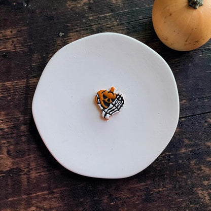 Finished orange jack-o-lantern wrapped in a white skeleton hand centered on a white round surface against a dark rustic wooden background with a pumpkin in the upper right.