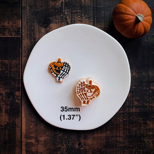 Finished orange jack-o-lantern and white skeleton hand clay piece beside the matching peach-colored cutter on a white round surface, with text showing 35mm (1.37").