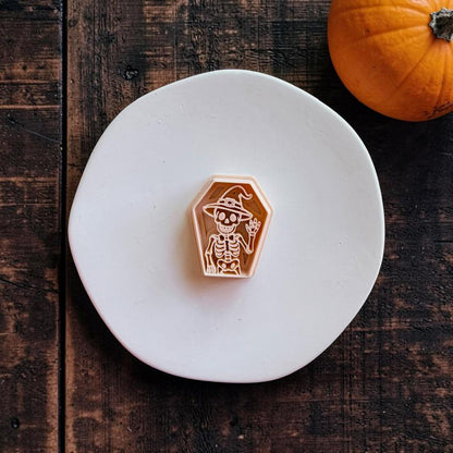 Peach-colored coffin-shaped polymer clay cutter featuring a waving skeleton wearing a pointed witch hat, centered on a white round surface with an orange pumpkin in the upper right.