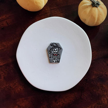 Finished gray coffin-shaped clay piece featuring a waving white skeleton in a witch hat with a small red bow accent, centered on a white round surface with pumpkins in the background.