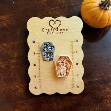Finished gray skeleton witch coffin clay piece beside the matching peach-colored polymer clay cutter on a CraftNLove Designs wooden measurement card.