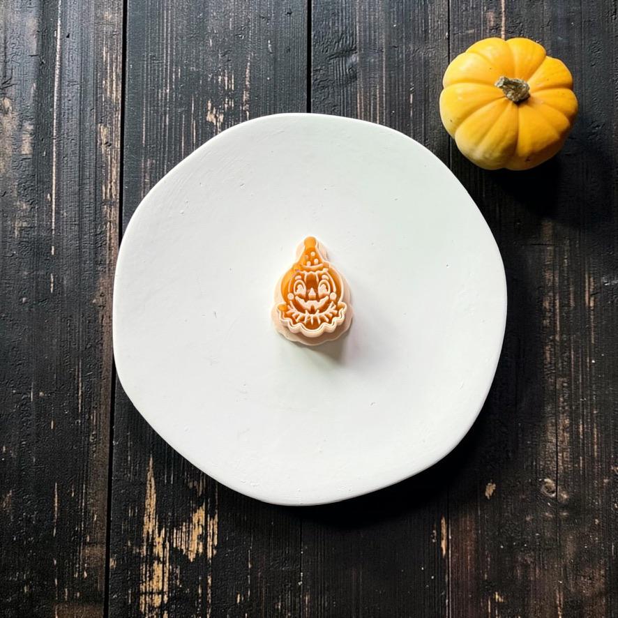 Peach-colored vintage clown jack-o-lantern design featuring a pointed hat, smiling pumpkin face, and ruffled collar, centered on a white round surface with a small orange pumpkin nearby.
