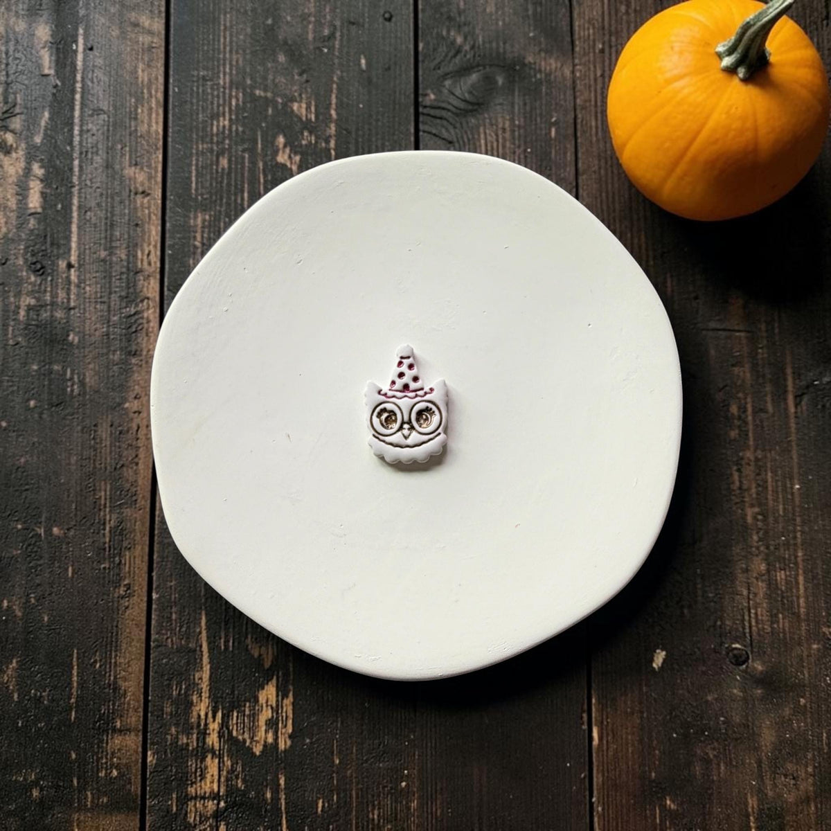 Finished white vintage-style owl with large round eyes and a pointed polka-dot witch hat centered on a white round surface, with an orange pumpkin in the upper right.
