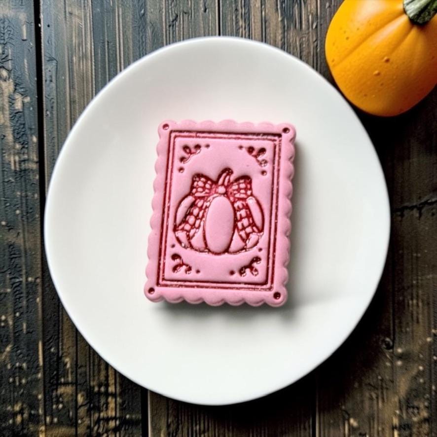 Finished pink rectangular clay piece with scalloped edges featuring a framed pumpkin wrapped with a large bow, small decorative corner flourishes, and four tiny corner holes, displayed on a white round surface.