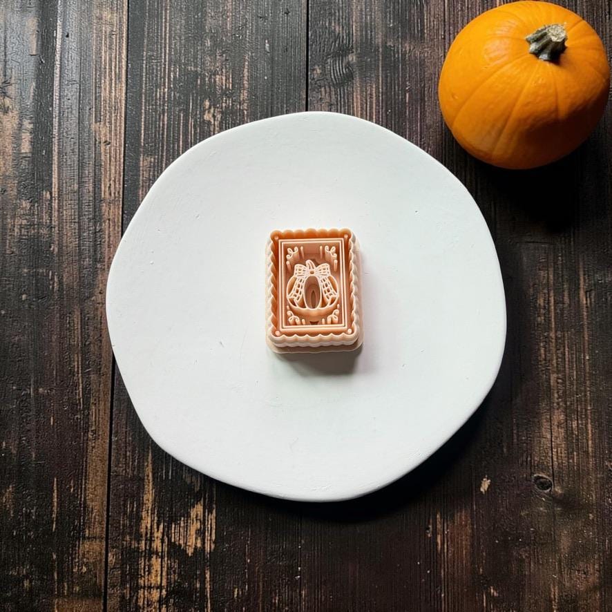 Peach-colored rectangular design with scalloped edges, an inset frame, decorative corner flourishes, and a pumpkin topped with a large bow, centered on a white round surface with an orange pumpkin nearby.