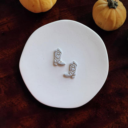 Finished mirrored ghost cowboy boot clay pieces displayed on a white round surface with small pumpkins in the background.