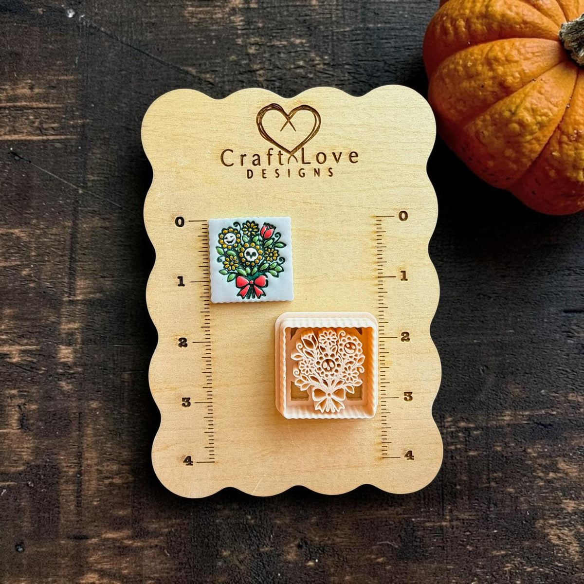 Finished colorful spooky bouquet piece and matching peach floral cutter displayed on a wooden Craft Love Designs measuring board with a pumpkin beside it.