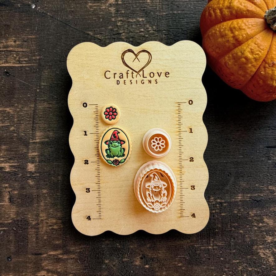 Finished colorful witchy frog and flower pieces shown beside their matching peach-colored designs on a wooden Craft Love Designs measuring board, with an orange pumpkin in the background