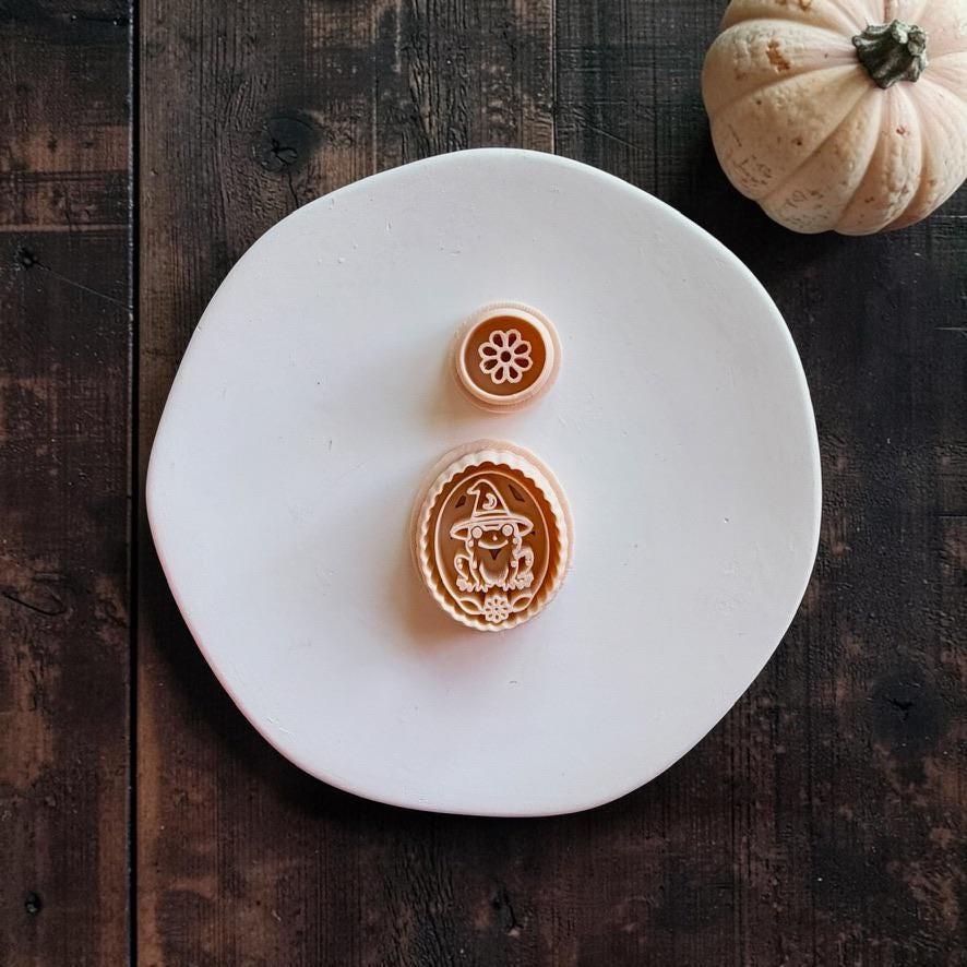 Peach-colored witchy frog and flower clay designs displayed on a white round surface. The oval frog design features a frog wearing a pointed witch hat with a small flower below, alongside a separate round flower design.