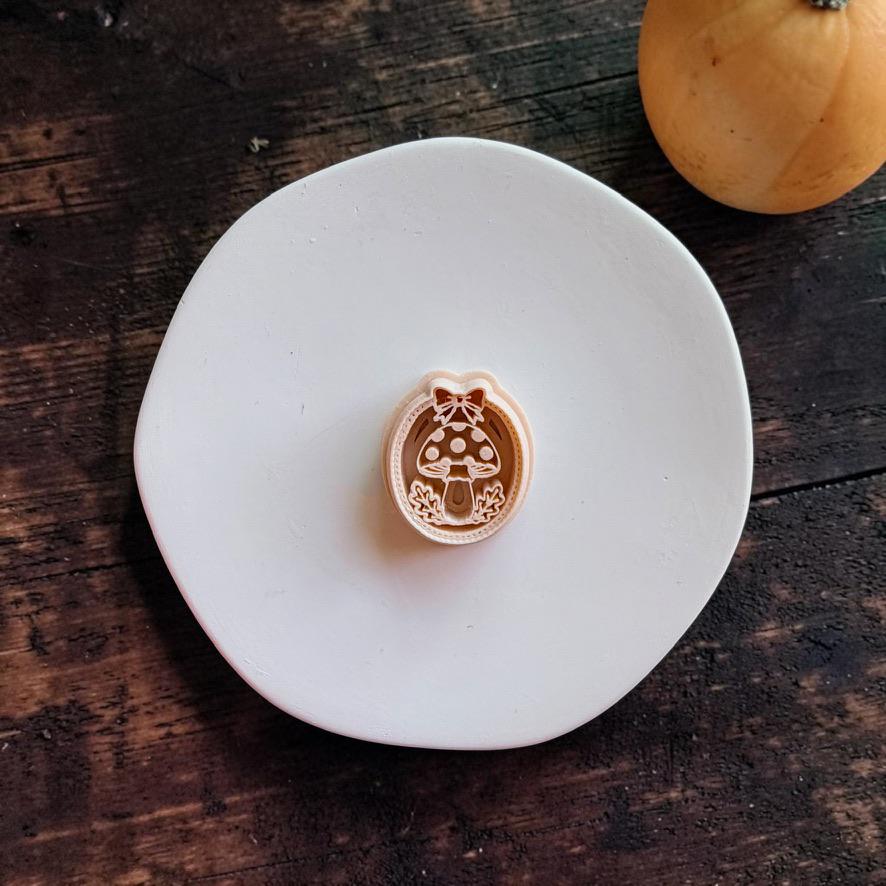 Peach-colored oval mushroom design centered on a white round surface. The design features a spotted toadstool with leafy accents, an oval decorative border, and a bow at the top, with a pumpkin in the background.