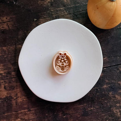 Peach-colored oval mushroom design centered on a white round surface. The design features a spotted toadstool with leafy accents, an oval decorative border, and a bow at the top, with a pumpkin in the background.