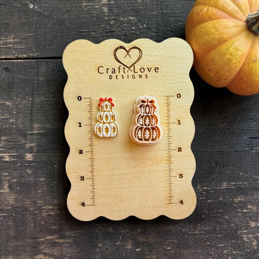 Finished white and gold stacked pumpkin clay piece with a red bow displayed beside the matching peach-colored design on a Craft Love Designs wooden measuring board. Three pumpkins are stacked vertically and decorated with leaf motifs.