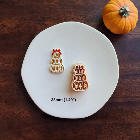 Finished white stacked pumpkin piece with golden-brown leaf details and a red bow displayed beside the larger peach-colored design on a white round surface. Text shows the size as 38mm (1.49 inches).