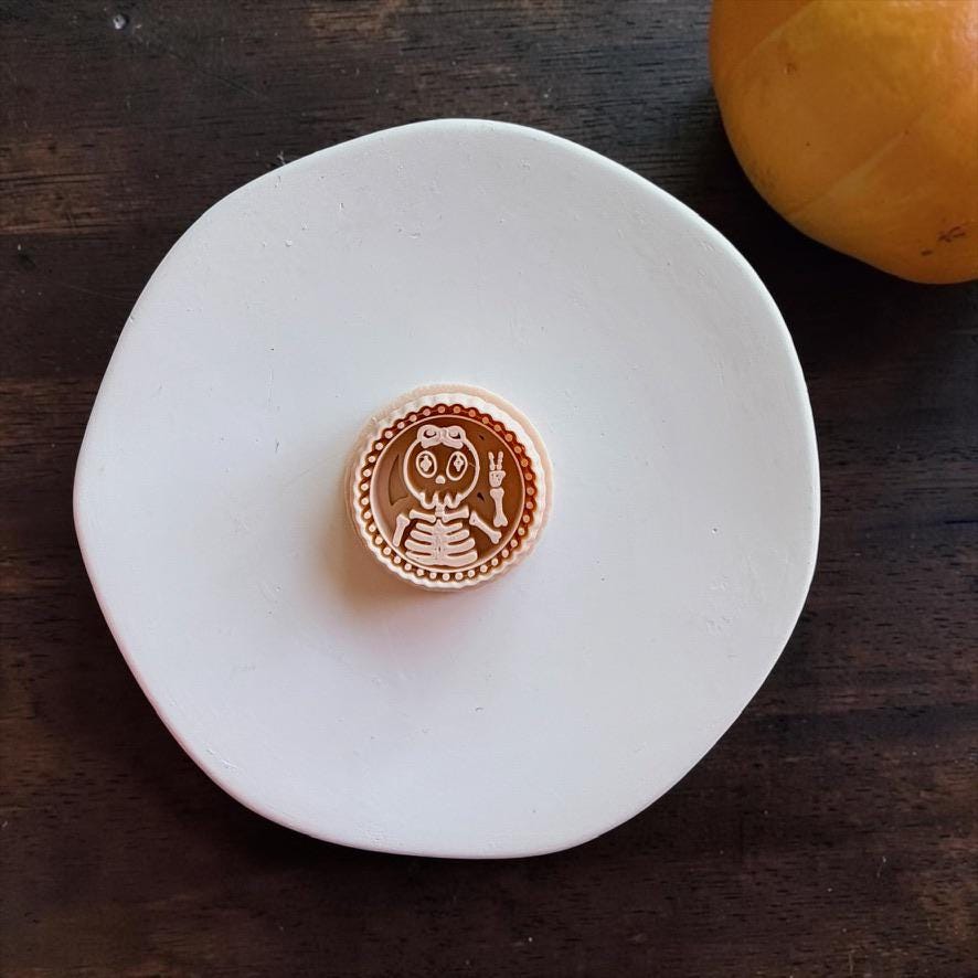 Peach-colored skeleton girl design centered on a white round surface, featuring flowing hair, large eyes, visible ribs, a raised peace sign hand, and a round beaded border.