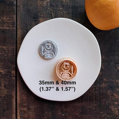 Gray and peach skeleton girl examples displayed together on a white round surface. The design features a skeleton girl making a peace sign, with text showing 35mm and 40mm sizes.