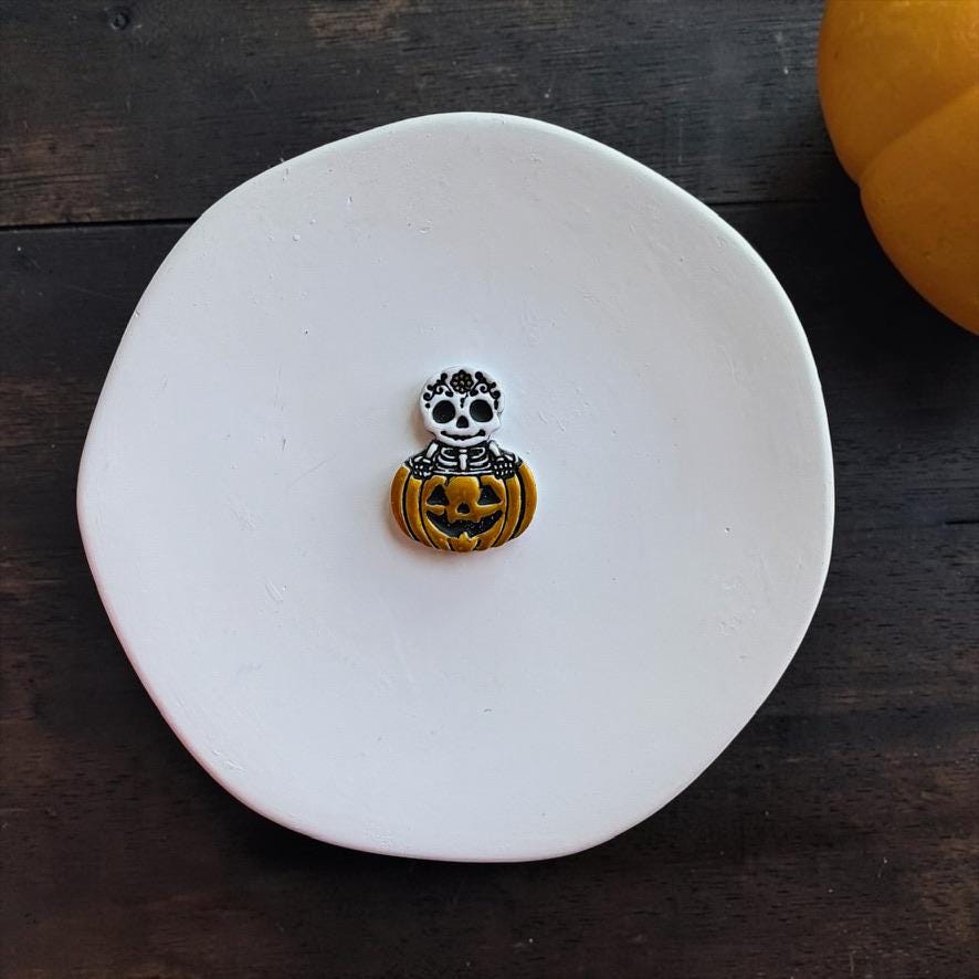 Finished skeleton pumpkin clay piece centered on a white round surface, with a white decorative skull and rib cage emerging from an orange jack-o-lantern.