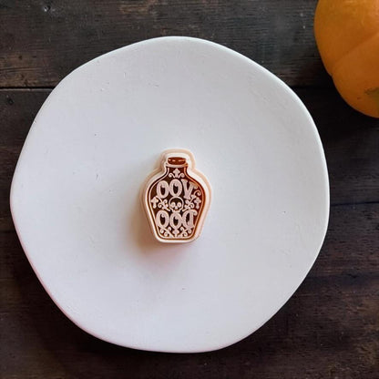 Peach-colored potion bottle design centered on a white round surface, featuring decorative lettering, a small skull, gothic flourishes, and an outlined bottle shape.