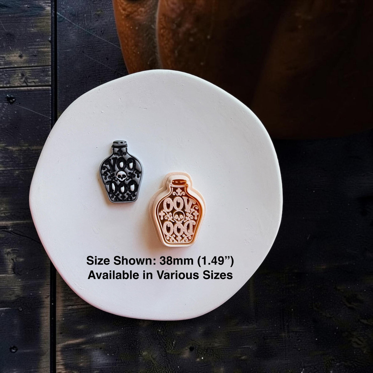 Black and gray finished skull potion bottle beside the larger peach-colored version on a white round surface. Text states size shown is 38mm (1.49") and available in various sizes.
