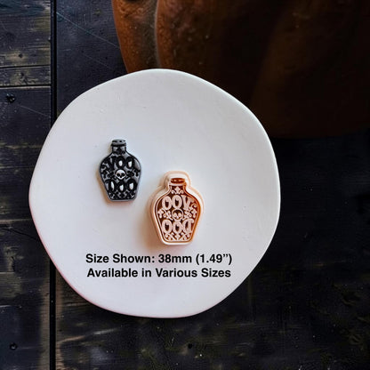 Black and gray finished skull potion bottle beside the larger peach-colored version on a white round surface. Text states size shown is 38mm (1.49") and available in various sizes.