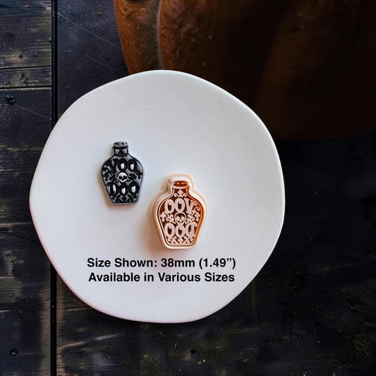 Black and gray finished skull potion bottle beside the larger peach-colored version on a white round surface. Text states size shown is 38mm (1.49") and available in various sizes.