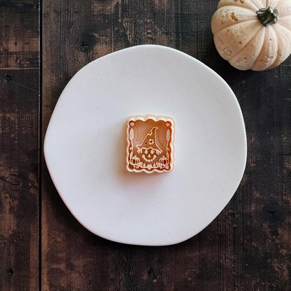 Peach-colored rectangular design centered on a white round surface, featuring a witchy scarecrow wearing a floppy pointed hat with autumn details and a decorative scalloped frame.