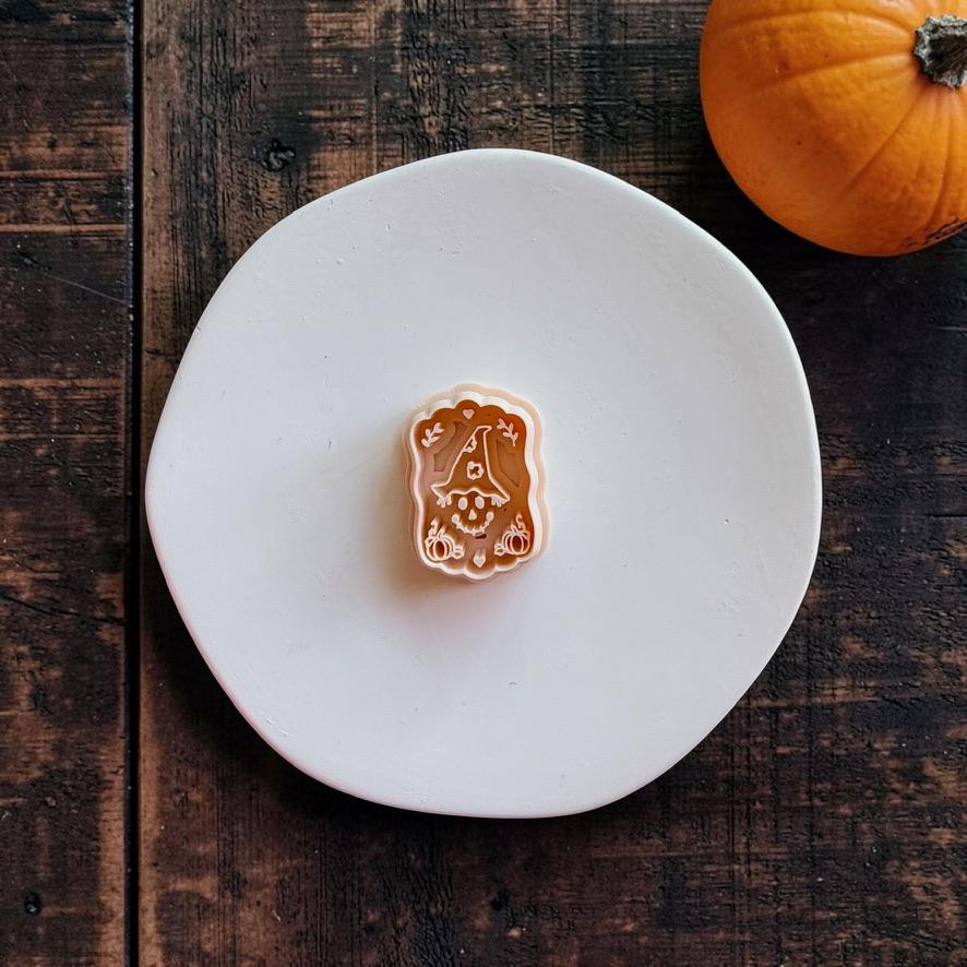 Peach-colored witchy scarecrow design centered on a white round surface, featuring an oversized pointed witch hat, stitched face, leaves, pumpkins, hearts, and a scalloped outer shape.