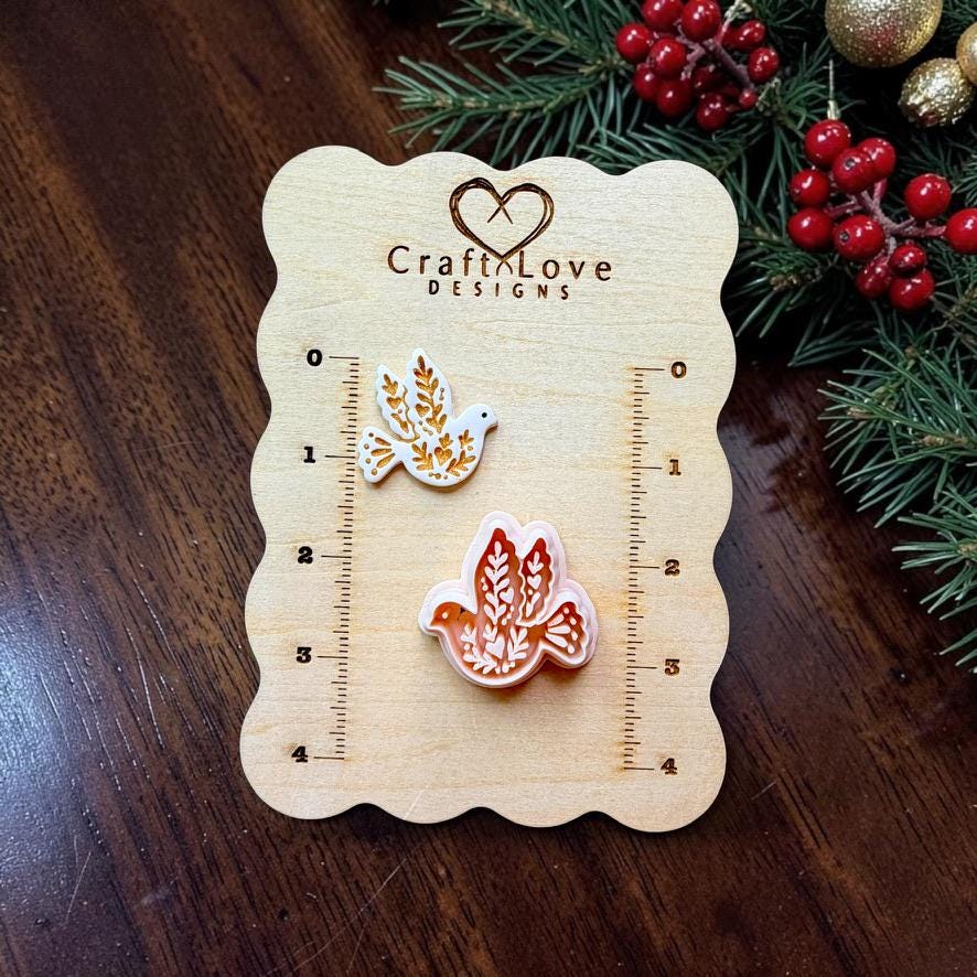 White and gold finished botanical dove beside the larger peach-colored dove design on a Craft Love Designs wooden measuring board, surrounded by evergreen branches, red berries, and gold Christmas ornaments.