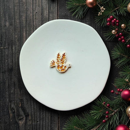 White and metallic gold finished folk-art dove centered on a white round surface, showing the leafy botanical pattern across its wings and body, with Christmas evergreen branches, berries, and ornaments around the scene.