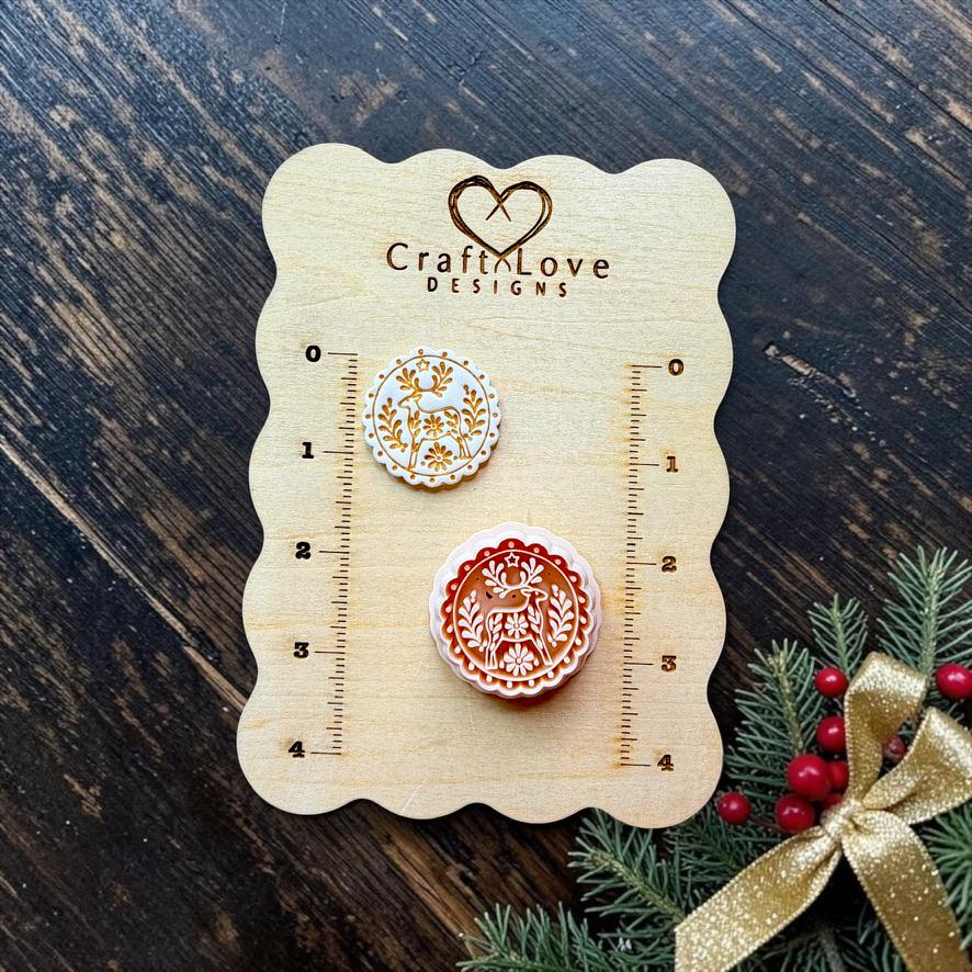 wo finished round folk art reindeer designs displayed on a Craft Love Designs wooden measuring board. The smaller white and gold version and larger red and white version feature a deer surrounded by botanical and floral details, with Christmas greenery and a gold bow nearby.