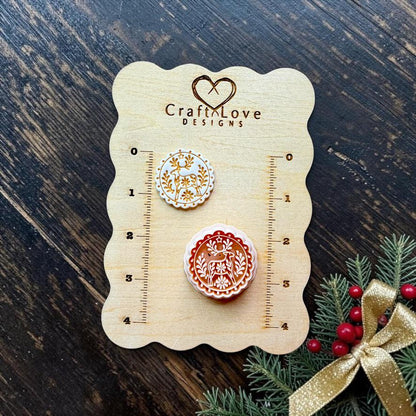 wo finished round folk art reindeer designs displayed on a Craft Love Designs wooden measuring board. The smaller white and gold version and larger red and white version feature a deer surrounded by botanical and floral details, with Christmas greenery and a gold bow nearby.