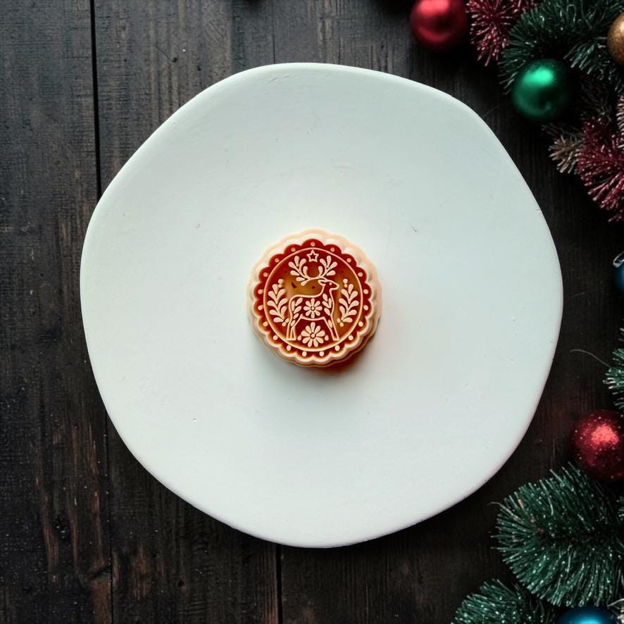 Large red, white, and gold round Christmas reindeer design centered on a white surface, featuring a folk art deer, flowers, leafy branches, star detail, and decorative scalloped border with Christmas ornaments along the edge.