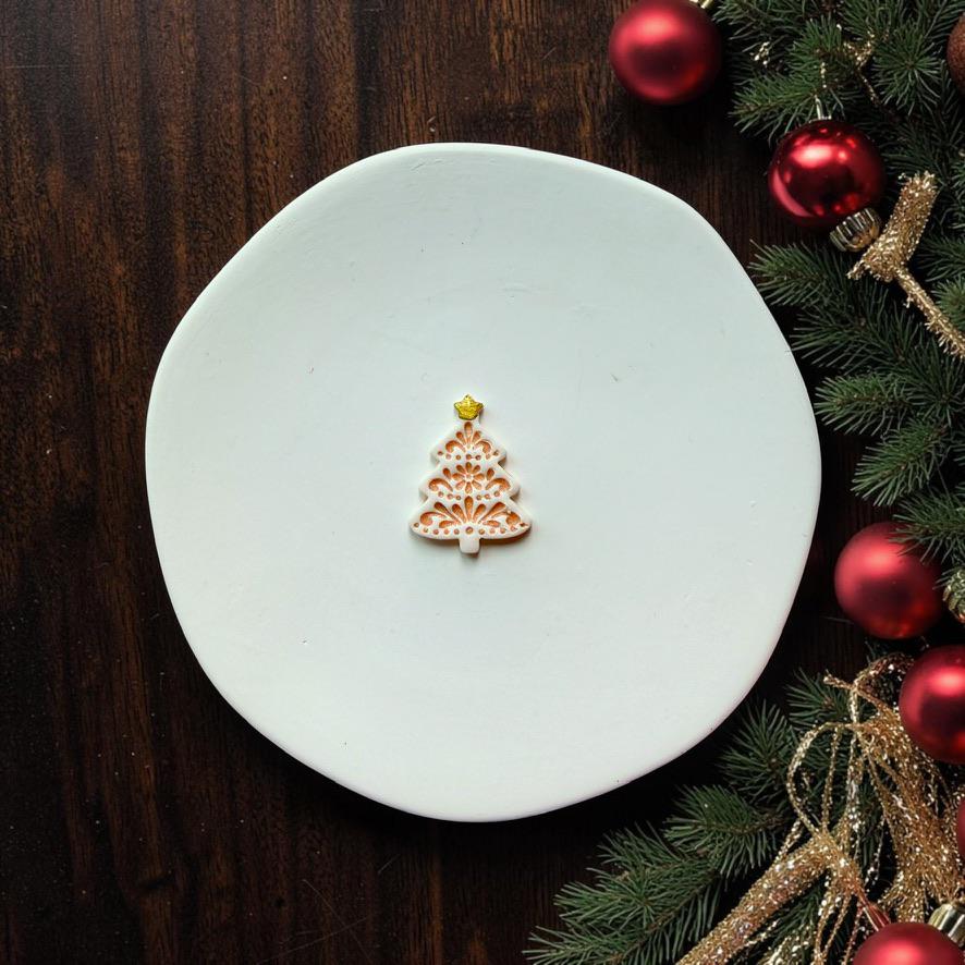 Small white and metallic gold folk art Christmas tree design centered on a white surface with red and gold Christmas ornaments and evergreen branches along the side.