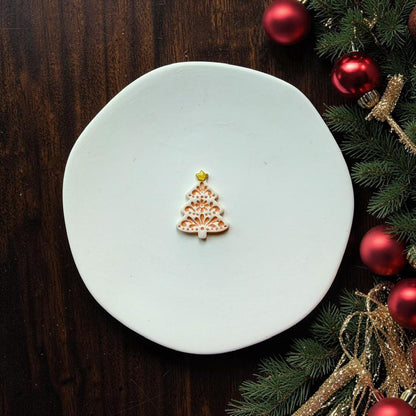 Small white and metallic gold folk art Christmas tree design centered on a white surface with red and gold Christmas ornaments and evergreen branches along the side.
