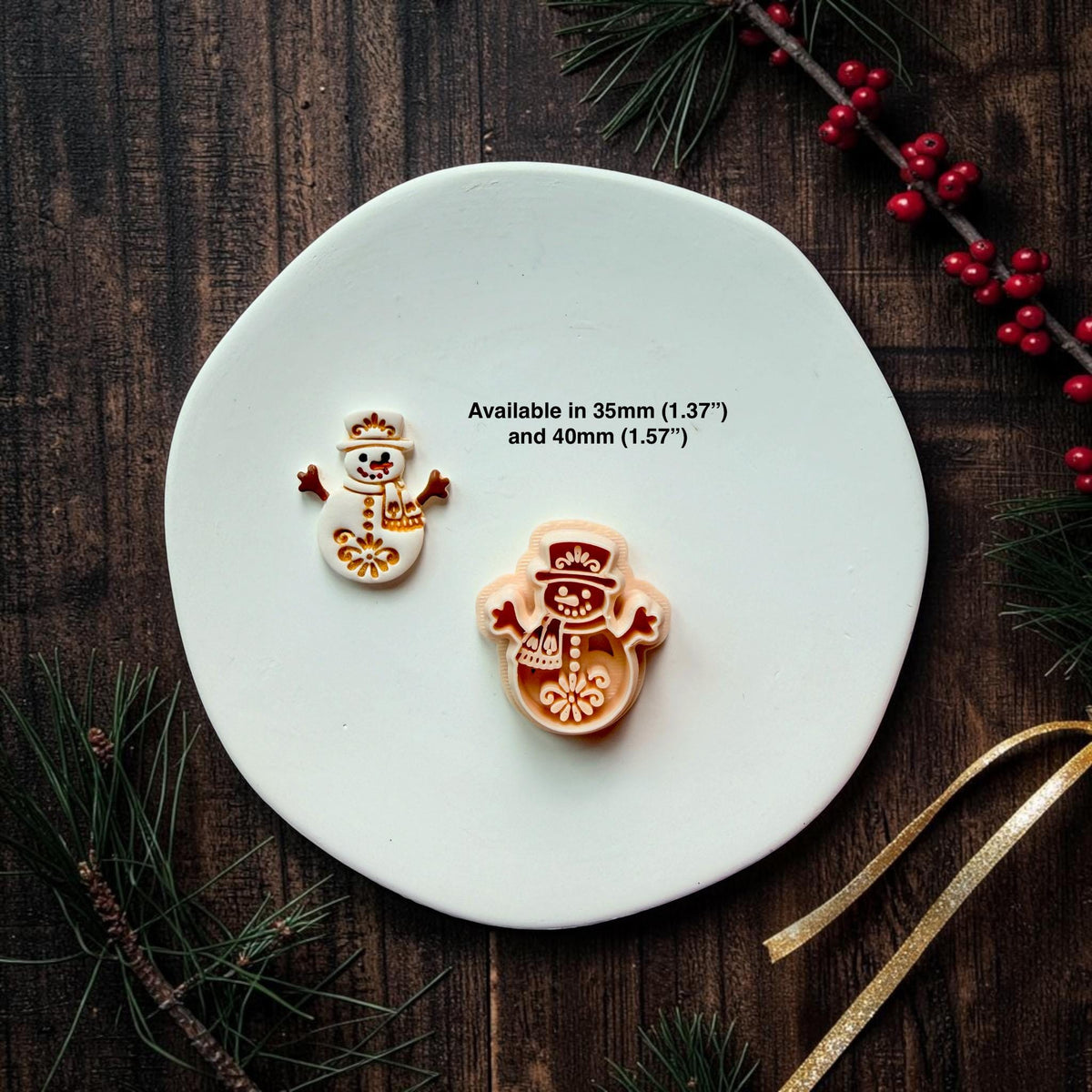 Small white and gold snowman beside a larger red and cream version on a white surface. Both feature top hats, scarves, raised arms, and decorative folk art details. Text states available in 35mm (1.37") and 40mm (1.57").