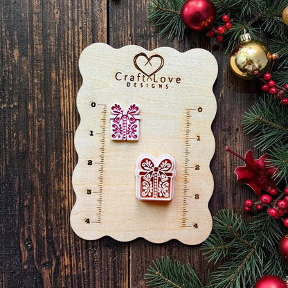 Small and large folk art Christmas present designs displayed on a Craft Love Designs wooden measuring board. Each gift box features a large bow, ribbon, floral details, and decorative dots, with evergreen branches, red berries, and Christmas ornaments nearby.