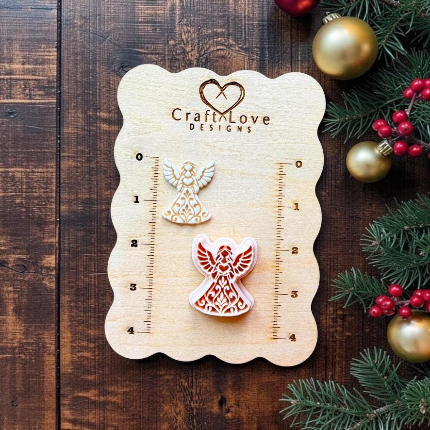 Two sizes of ornate Christmas angel designs displayed on a Craft Love Designs wooden measuring board. The angels have raised wings, flowing gowns, and detailed folk art scrollwork, surrounded by evergreen branches, red berries, and gold Christmas ornaments.