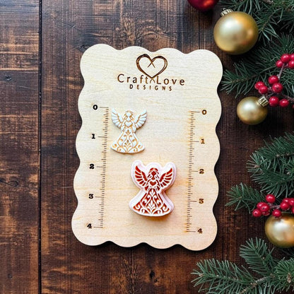Two sizes of ornate Christmas angel designs displayed on a Craft Love Designs wooden measuring board. The angels have raised wings, flowing gowns, and detailed folk art scrollwork, surrounded by evergreen branches, red berries, and gold Christmas ornaments.