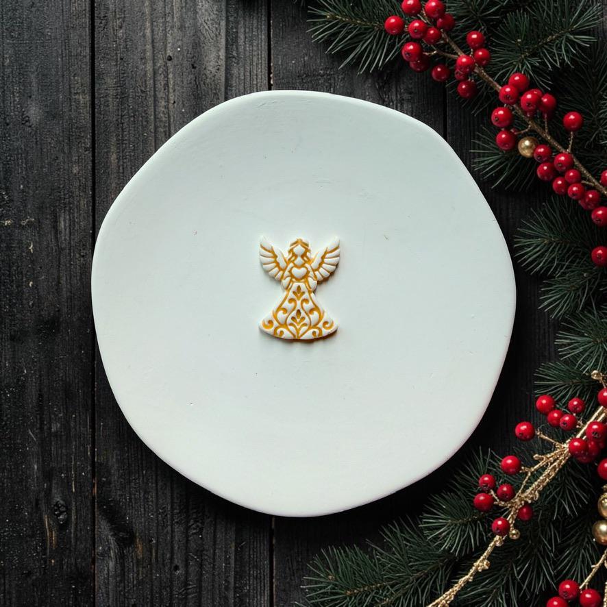 Small white and gold Christmas angel design centered on a white surface, featuring detailed wings and an ornate flowing gown, with evergreen branches and red berries along the edge.