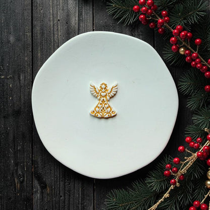 Small white and gold Christmas angel design centered on a white surface, featuring detailed wings and an ornate flowing gown, with evergreen branches and red berries along the edge.