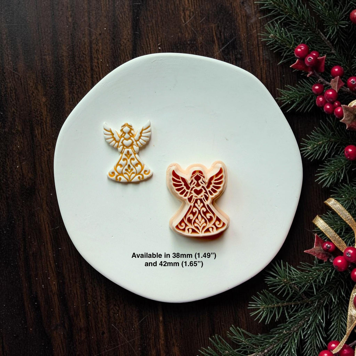 Small white and gold angel beside a larger red and cream angel on a white surface with Christmas greenery and red berries. Both feature raised wings and decorative folk art gowns. Available in 38mm (1.49") and 42mm (1.65").