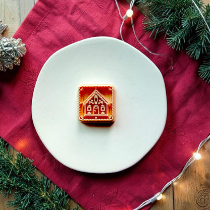 Larger brown and cream gingerbread house design centered on a white plate over a red holiday cloth, surrounded by evergreen branches, Christmas decorations, and glowing string lights.