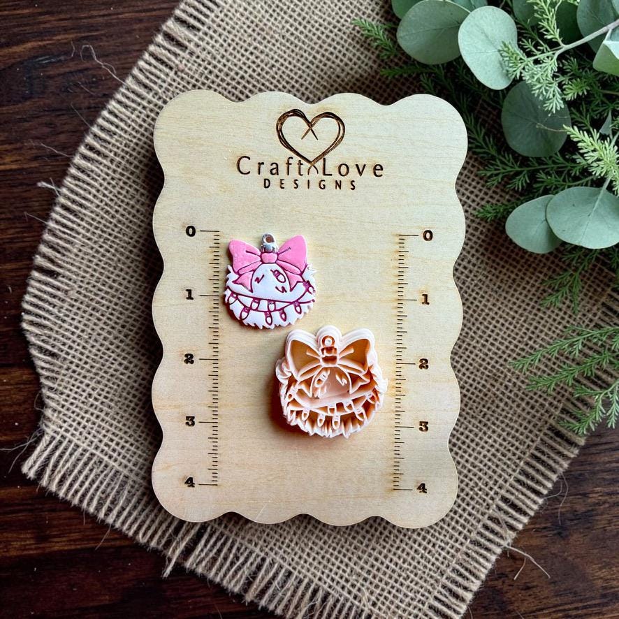 Finished pink and white mischievous Christmas ornament design beside the matching clay cutter on a Craft Love Designs wooden measurement board, displayed over burlap with greenery.