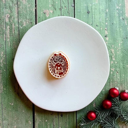 Matching reindeer clay cutter centered on a white plate against rustic green wood, showing the oval frame, antlers, reindeer face, scarf, and bow details, with evergreen and red ornaments in the corner.
