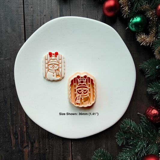 Finished white snowman design centered on a white plate, featuring engraved details, a red bow, winter hat, smiling face, scarf, and buttons, with Christmas greenery and ornaments in the background.