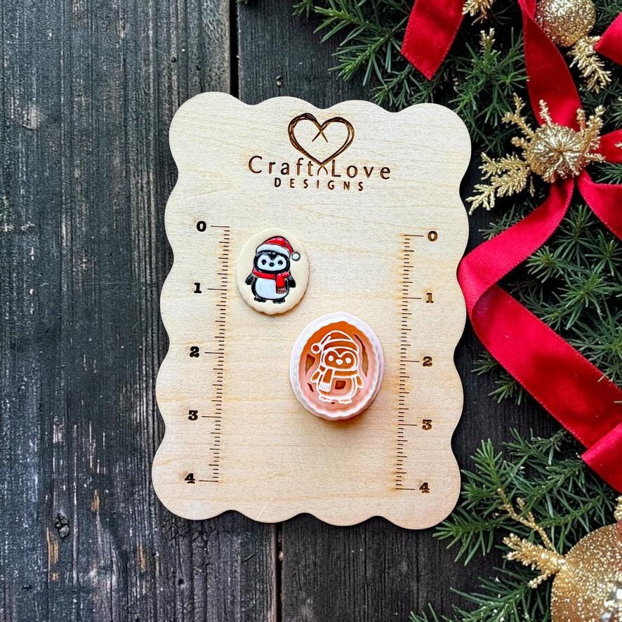 Finished Christmas penguin clay piece and matching larger design displayed on a Craft Love Designs wooden measurement board, with the penguin wearing a red Santa hat and scarf beside festive greenery and red ribbon.