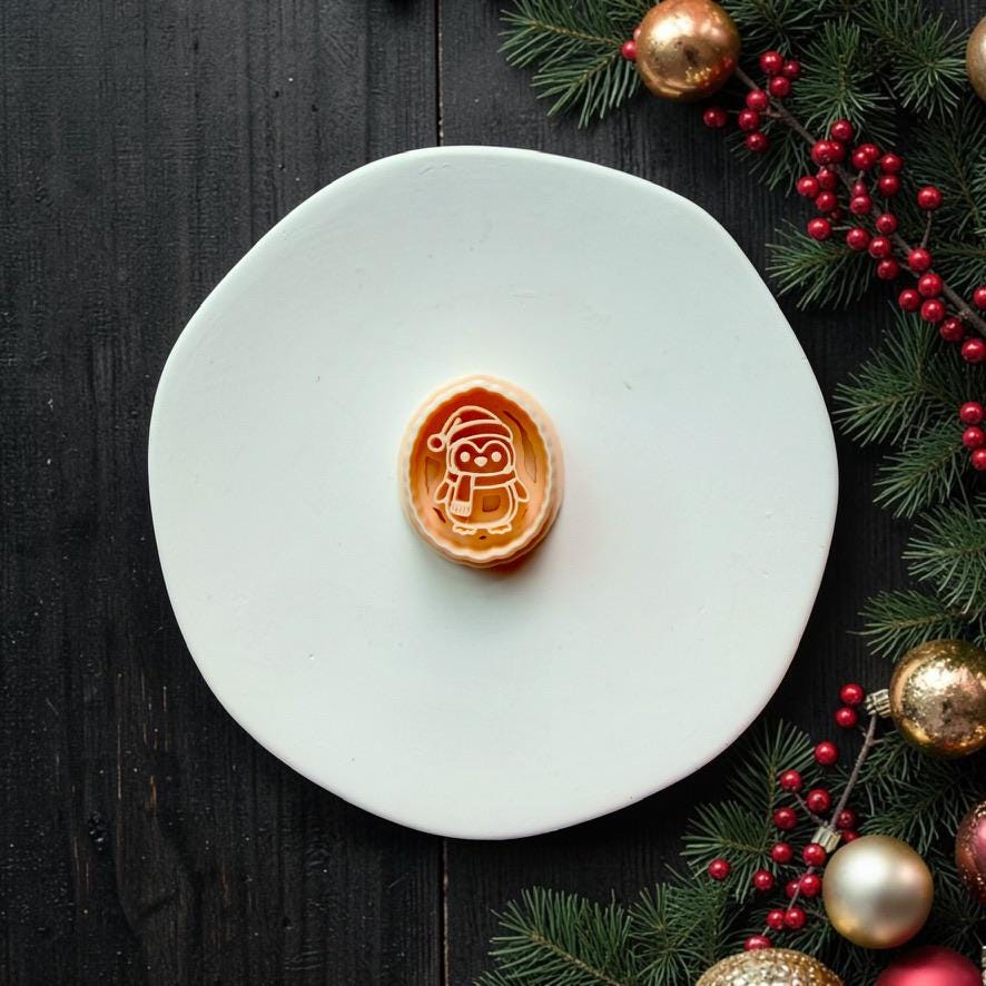 Oval Christmas penguin design centered on a white plate, showing a penguin wearing a Santa hat and scarf, with evergreen branches, red berries, and gold ornaments along the side.