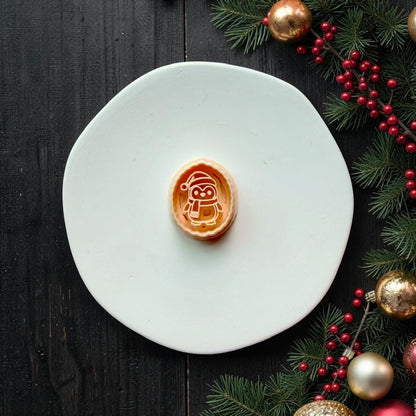 Oval Christmas penguin design centered on a white plate, showing a penguin wearing a Santa hat and scarf, with evergreen branches, red berries, and gold ornaments along the side.