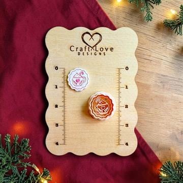Hot cocoa design displayed on a Craft Love Designs wooden measurement board, with a finished white clay piece above the matching orange design. Both feature a scalloped border and festive cocoa mug with heart and candy cane.