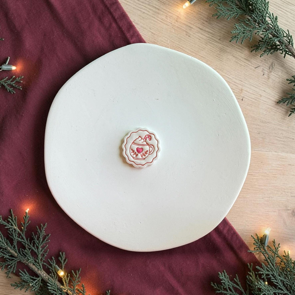 Finished white hot cocoa clay piece centered on a white plate, featuring red details including a scalloped border, heart-decorated mug, whipped topping, leaves, and candy cane, surrounded by greenery and warm Christmas lights.
