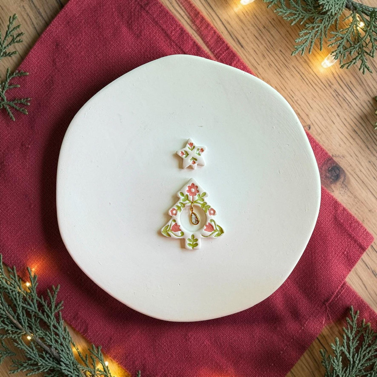 Finished folk art Christmas tree earring centered on a white plate. The white clay tree has pink and green floral details, a coordinating floral star topper, and a gold teardrop charm hanging inside the circular tree opening.